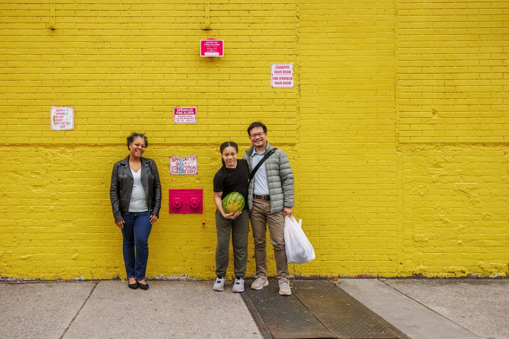 Family of three standing together outdoors against a bright yellow wall